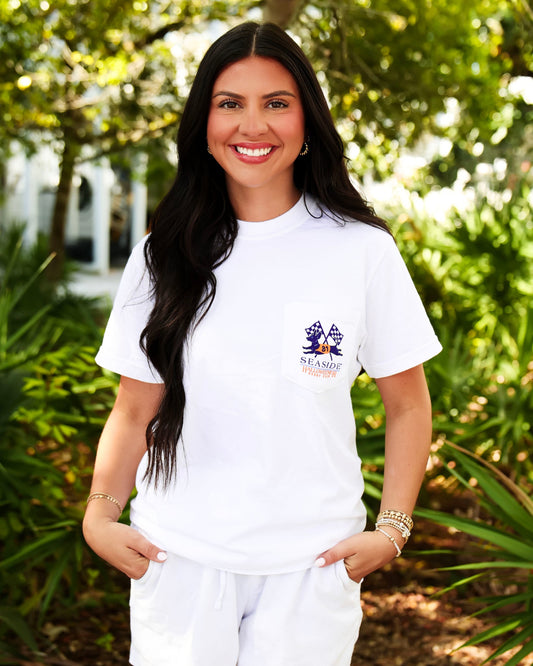 A smiling woman with long dark hair stands outdoors wearing a white short-sleeve crewneck t-shirt with the halloweener derby logo on the left chest, paired with white shorts. The background is a lush, sunlit garden with green foliage.