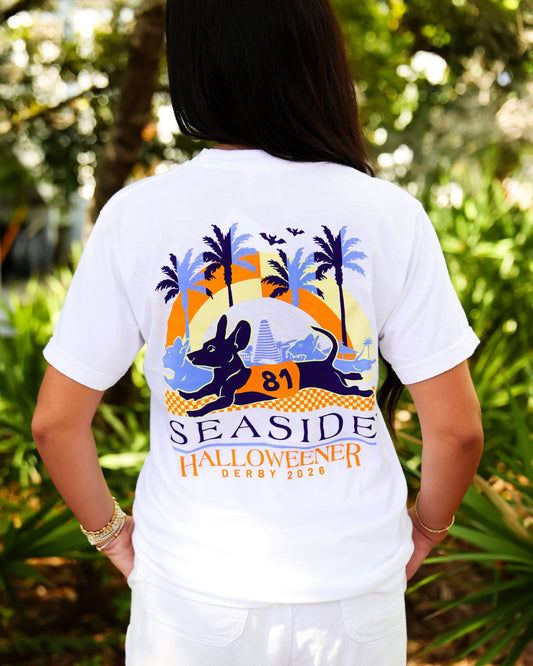 A woman viewed from behind wearing a white graphic T-shirt outdoors. The back of the shirt features a large seaside halloweener derby graphic.