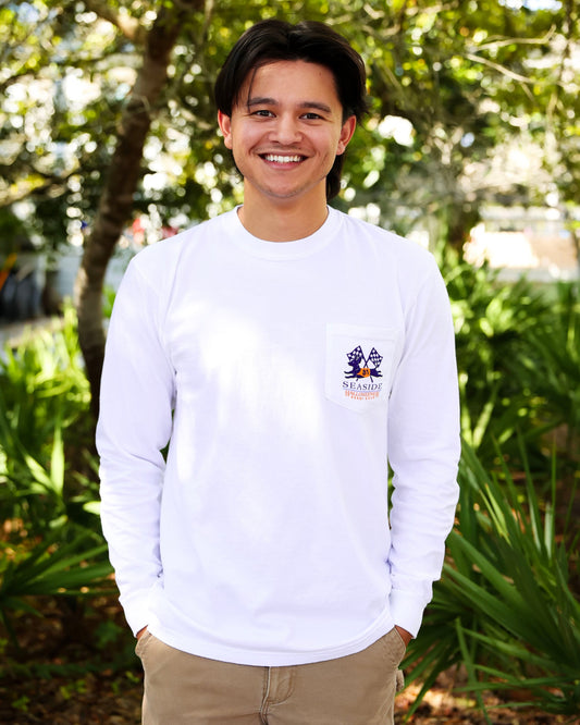 A smiling man wearing a white long-sleeve t-shirt with a small Seaside Halloweener Derby graphic, standing outdoors in front of a lush green background of trees and plants.