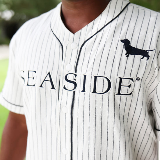 A close-up of a person wearing a white baseball jersey with thin black pinstripes, black piping, and a black dachshund logo on the chest. The word 'SEASIDE' is printed across the front.