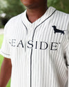 A close-up of a person wearing a white baseball jersey with thin black pinstripes, black piping, and a black dachshund logo on the chest. The word 'SEASIDE' is printed across the front.