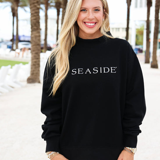 A smiling blonde woman wearing a black crewneck sweatshirt with 'SEASIDE' printed in white across the chest, paired with white shorts, standing outdoors on a sunny beachside walkway with palm trees in the background.