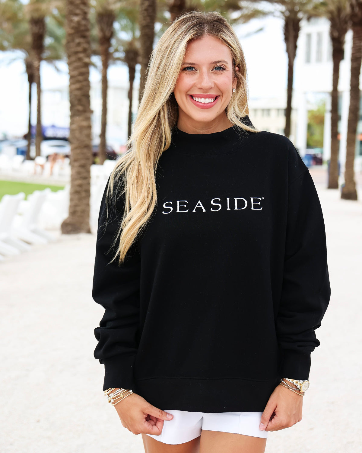 A smiling blonde woman wearing a black crewneck sweatshirt with 'SEASIDE' printed in white across the chest, paired with white shorts, standing outdoors on a sunny beachside walkway with palm trees in the background.