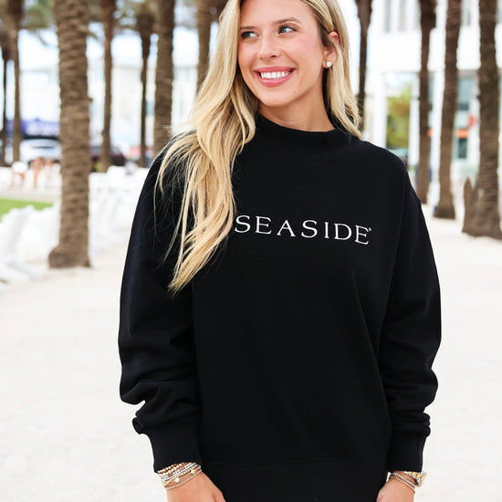 A woman wearing a black crewneck sweatshirt with 'SEASIDE' printed in white across the chest, standing outdoors on a sandy path lined with palm trees.