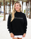 A woman wearing a black crewneck sweatshirt with 'SEASIDE' printed in white across the chest, standing outdoors on a sandy path lined with palm trees.