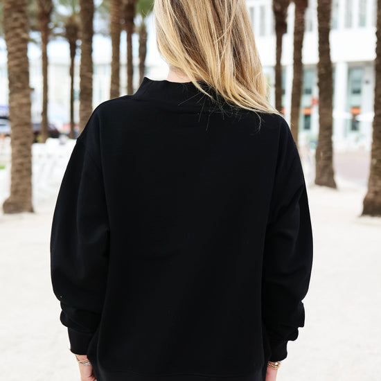 A woman viewed from behind wearing a black long-sleeve mock neck sweatshirt and white shorts, standing outdoors on a sandy path lined with palm trees.