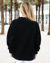A woman viewed from behind wearing a black long-sleeve mock neck sweatshirt and white shorts, standing outdoors on a sandy path lined with palm trees.