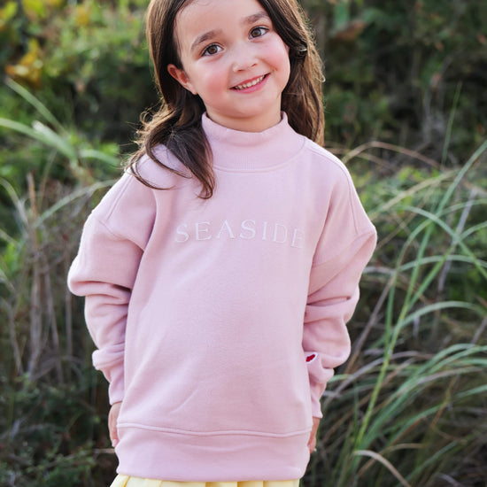 A young girl with brown hair smiling outdoors, wearing a light pink mock-neck sweatshirt with 'SEASIDE' printed monochromatic across the chest, paired with a yellow pleated skirt.