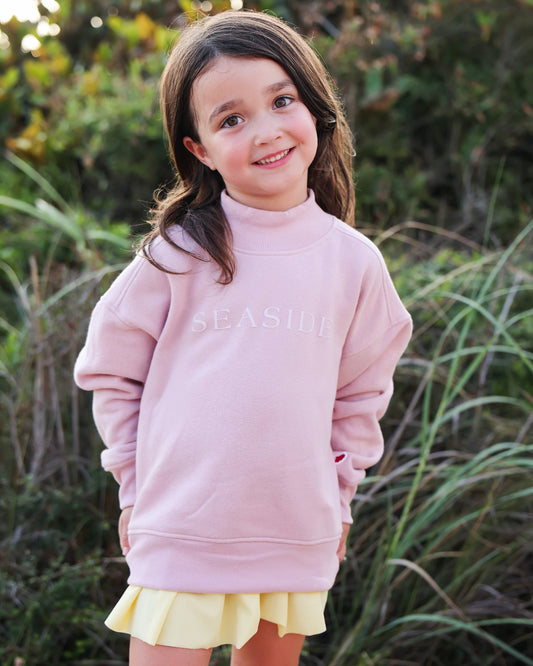 A young girl with brown hair smiling outdoors, wearing a light pink mock-neck sweatshirt with 'SEASIDE' printed monochromatic across the chest, paired with a yellow pleated skirt.