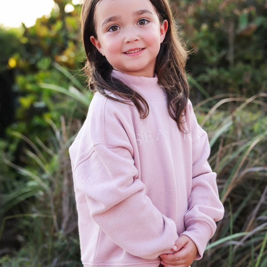 A young girl with long brown hair stands outdoors wearing a light pink sweatshirt and a yellow skirt. The background consists of soft-focus green foliage and grass.
