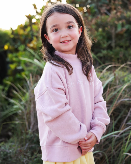 A young girl with long brown hair stands outdoors wearing a light pink sweatshirt and a yellow skirt. The background consists of soft-focus green foliage and grass.