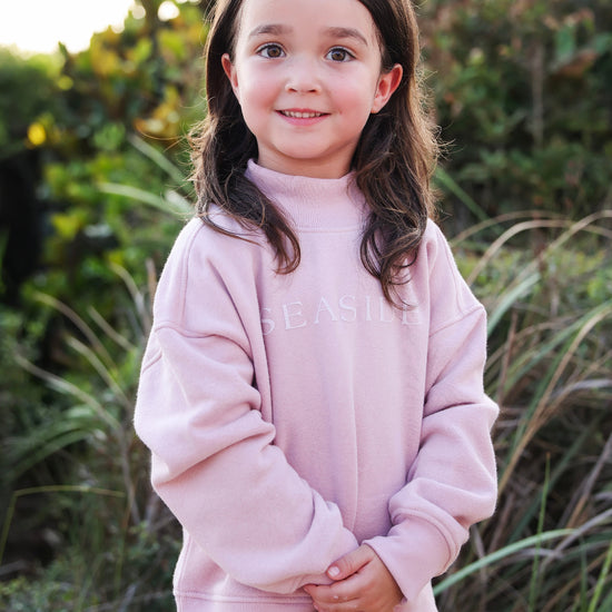 A young girl with long brown hair stands outdoors wearing a dusty pink sweatshirt with 'SEASIDE' printed across the chest and a yellow skirt, set against a blurred natural background of green foliage and grass.