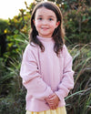 A young girl with long brown hair stands outdoors wearing a dusty pink sweatshirt with 'SEASIDE' printed across the chest and a yellow skirt, set against a blurred natural background of green foliage and grass.