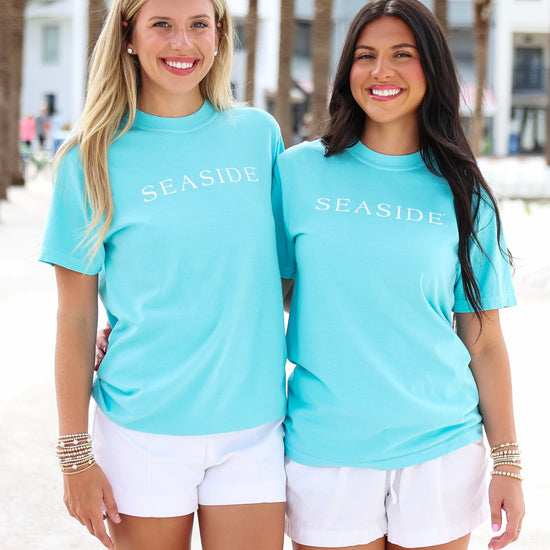 Two women standing on a beach wearing matching turquoise short-sleeve t-shirts with 'SEASIDE' printed in white across the chest, paired with white shorts.