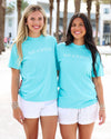 Two women standing on a beach wearing matching turquoise short-sleeve t-shirts with 'SEASIDE' printed in white across the chest, paired with white shorts.