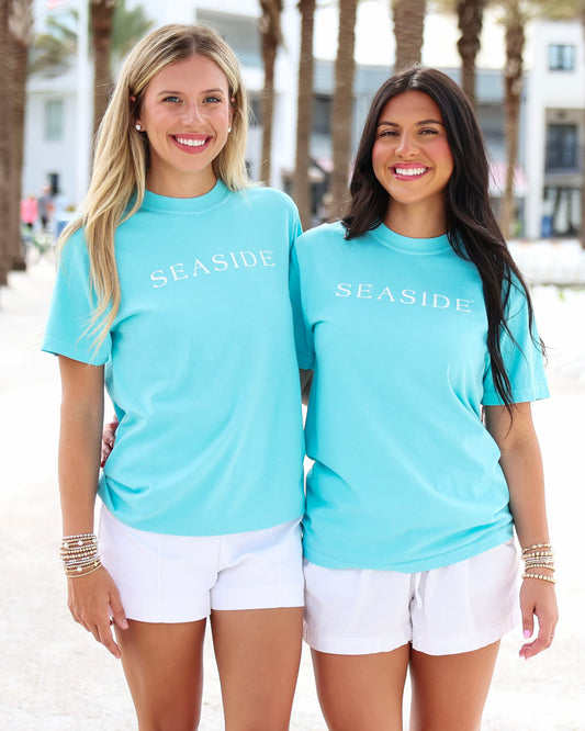 Two women standing on a beach wearing matching turquoise short-sleeve t-shirts with 'SEASIDE' printed in white across the chest, paired with white shorts.