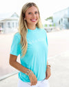 A smiling blonde woman wearing a bright turquoise short-sleeve t-shirt with 'SEASIDE' printed in white across the chest, paired with white pants, standing outdoors on a sunny day.