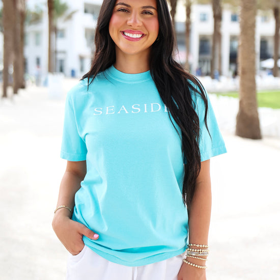 A woman wearing a light blue t-shirt with 'SEASIDE' printed in white across the chest, paired with white shorts, standing outdoors on a sunny beachside path with palm trees in the background.