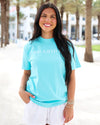A woman wearing a light blue t-shirt with 'SEASIDE' printed in white across the chest, paired with white shorts, standing outdoors on a sunny beachside path with palm trees in the background.