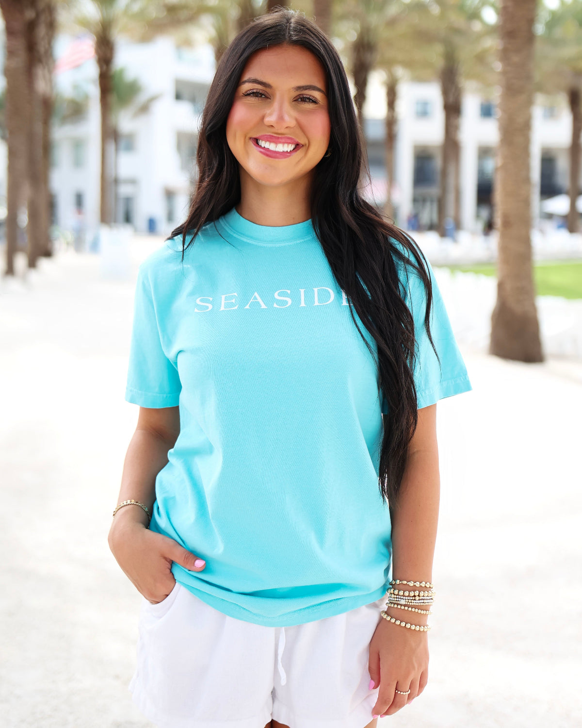 A woman wearing a light blue t-shirt with 'SEASIDE' printed in white across the chest, paired with white shorts, standing outdoors on a sunny beachside path with palm trees in the background.