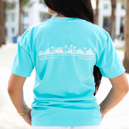 A woman viewed from behind wearing a light turquoise blue t-shirt with a white graphic of a beach town skyline and the text 'The New Town' and 'The Old Ways' printed across the back.