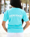 A woman viewed from behind wearing a light turquoise blue t-shirt with a white graphic of a beach town skyline and the text 'The New Town' and 'The Old Ways' printed across the back.