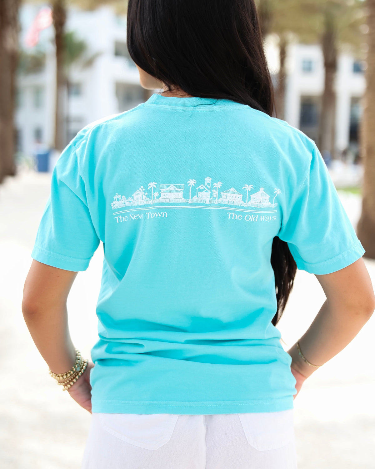 A woman viewed from behind wearing a light turquoise blue t-shirt with a white graphic of a beach town skyline and the text 'The New Town' and 'The Old Ways' printed across the back.