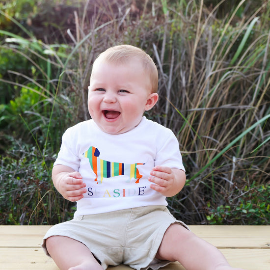 A smiling baby sitting outdoors on a wooden deck wearing a white t-shirt with a colorful dachshund graphic and the word 'SEASIDE', paired with khaki shorts.