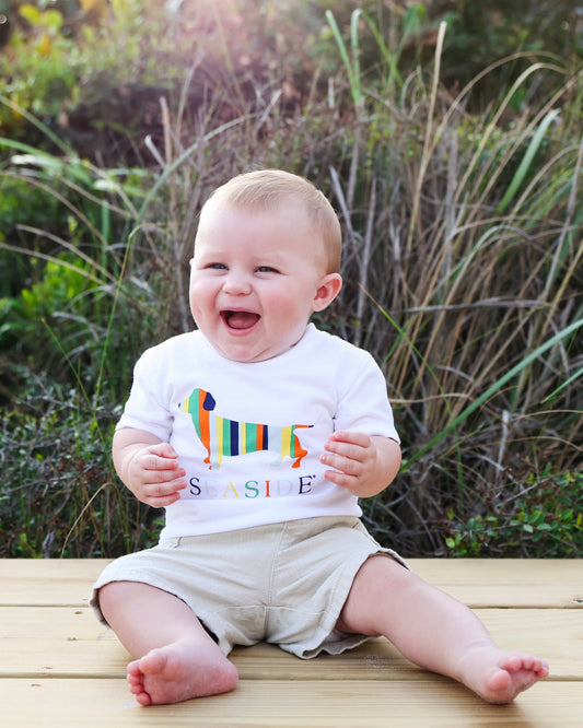 A smiling baby sitting outdoors on a wooden deck wearing a white t-shirt with a colorful dachshund graphic and the word 'SEASIDE', paired with khaki shorts.