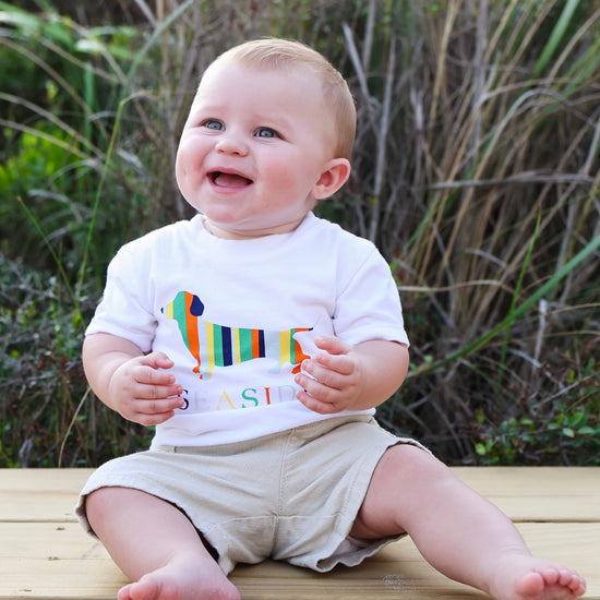 A baby sitting on a wooden deck outdoors, wearing a white t-shirt with a colorful striped dachshund graphic and the word 'SEASIDE' on the front, paired with beige shorts.