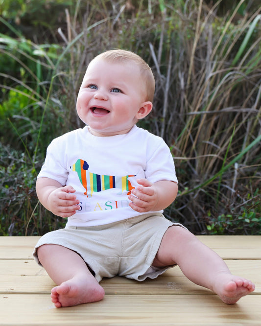 A baby sitting on a wooden deck outdoors, wearing a white t-shirt with a colorful striped dachshund graphic and the word 'SEASIDE' on the front, paired with beige shorts.