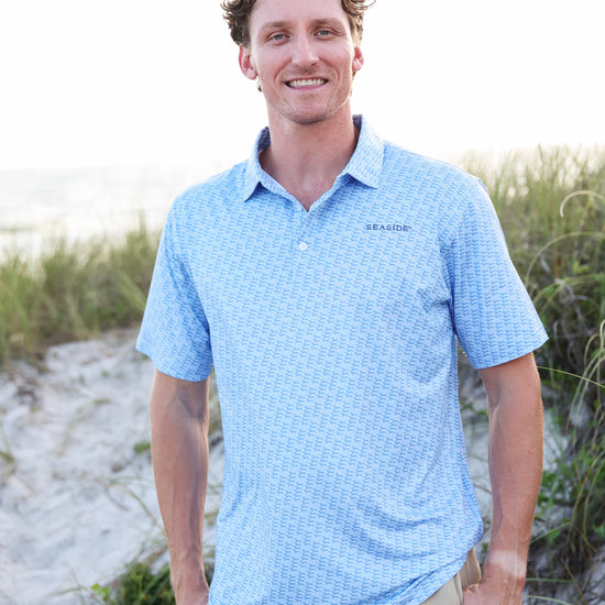A man standing on a sandy beach wearing a light blue dachshund patterned short-sleeve polo shirt and khaki pants.