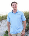 A man standing on a sandy beach wearing a light blue dachshund patterned short-sleeve polo shirt and khaki pants.