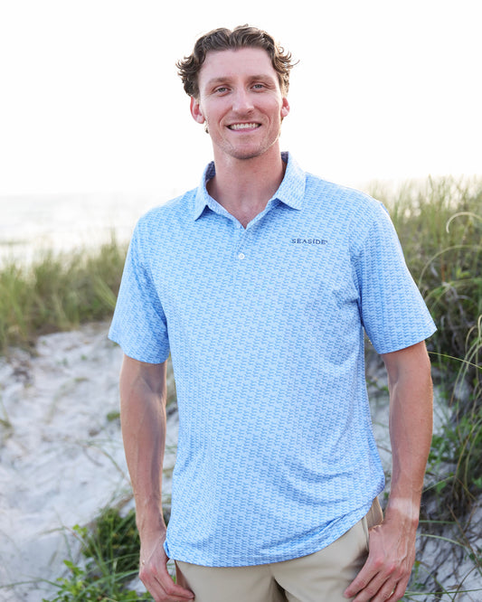 A man standing on a sandy beach wearing a light blue dachshund patterned short-sleeve polo shirt and khaki pants.