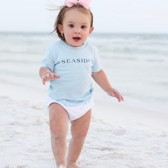 A toddler walking barefoot on a sandy beach wearing a light blue short-sleeve t-shirt with 'SEASIDE' printed across the chest, white diaper cover, and a pink bow headband.