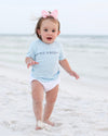 A toddler walking barefoot on a sandy beach wearing a light blue short-sleeve t-shirt with 'SEASIDE' printed across the chest, white diaper cover, and a pink bow headband.