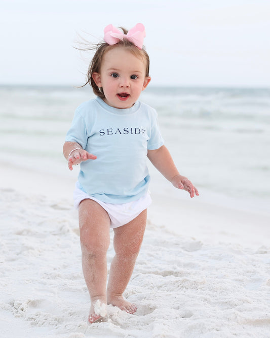 A toddler walking barefoot on a sandy beach wearing a light blue short-sleeve t-shirt with 'SEASIDE' printed across the chest, white diaper cover, and a pink bow headband.