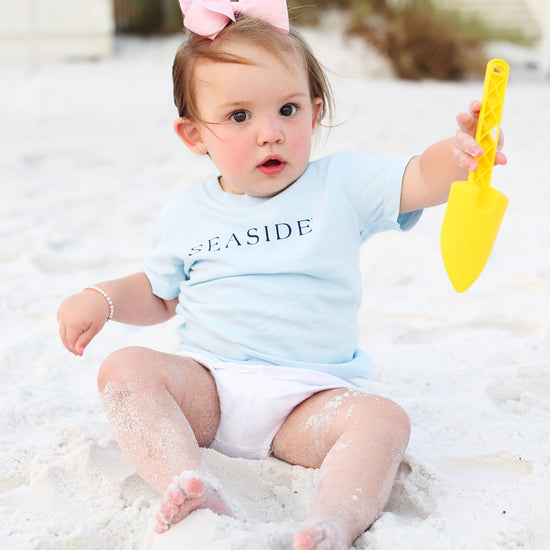 A baby girl sitting on white sand wearing a light blue short-sleeve t-shirt with 'SEASIDE' printed in navy text, white diaper cover, and a pink bow in her hair, while holding a yellow plastic toy shovel.
