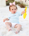 A baby girl sitting on white sand wearing a light blue short-sleeve t-shirt with 'SEASIDE' printed in navy text, white diaper cover, and a pink bow in her hair, while holding a yellow plastic toy shovel.