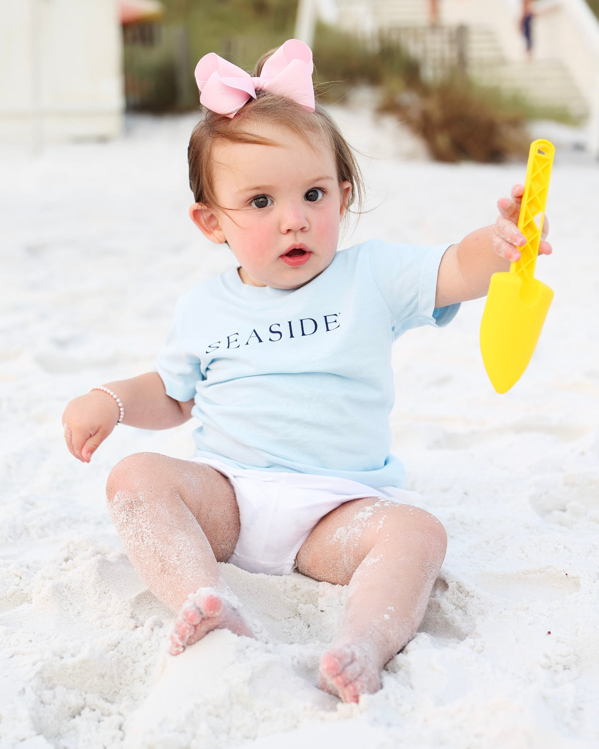 A baby girl sitting on white sand wearing a light blue short-sleeve t-shirt with 'SEASIDE' printed in navy text, white diaper cover, and a pink bow in her hair, while holding a yellow plastic toy shovel.