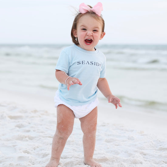 A toddler standing on a sandy beach wearing a light blue t-shirt with 'SEASIDE' printed on the chest, a white diaper, and a pink bow headband. The ocean and a pale sky are visible in the background.