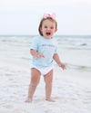 A toddler standing on a sandy beach wearing a light blue t-shirt with 'SEASIDE' printed on the chest, a white diaper, and a pink bow headband. The ocean and a pale sky are visible in the background.