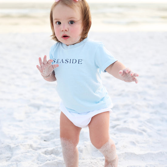 A toddler standing on a sandy beach wearing a light blue short-sleeve bodysuit with the word 'SEASIDE' printed in navy across the chest, along with a white diaper and a pink bow in her hair.