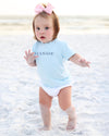A toddler standing on a sandy beach wearing a light blue short-sleeve bodysuit with the word 'SEASIDE' printed in navy across the chest, along with a white diaper and a pink bow in her hair.
