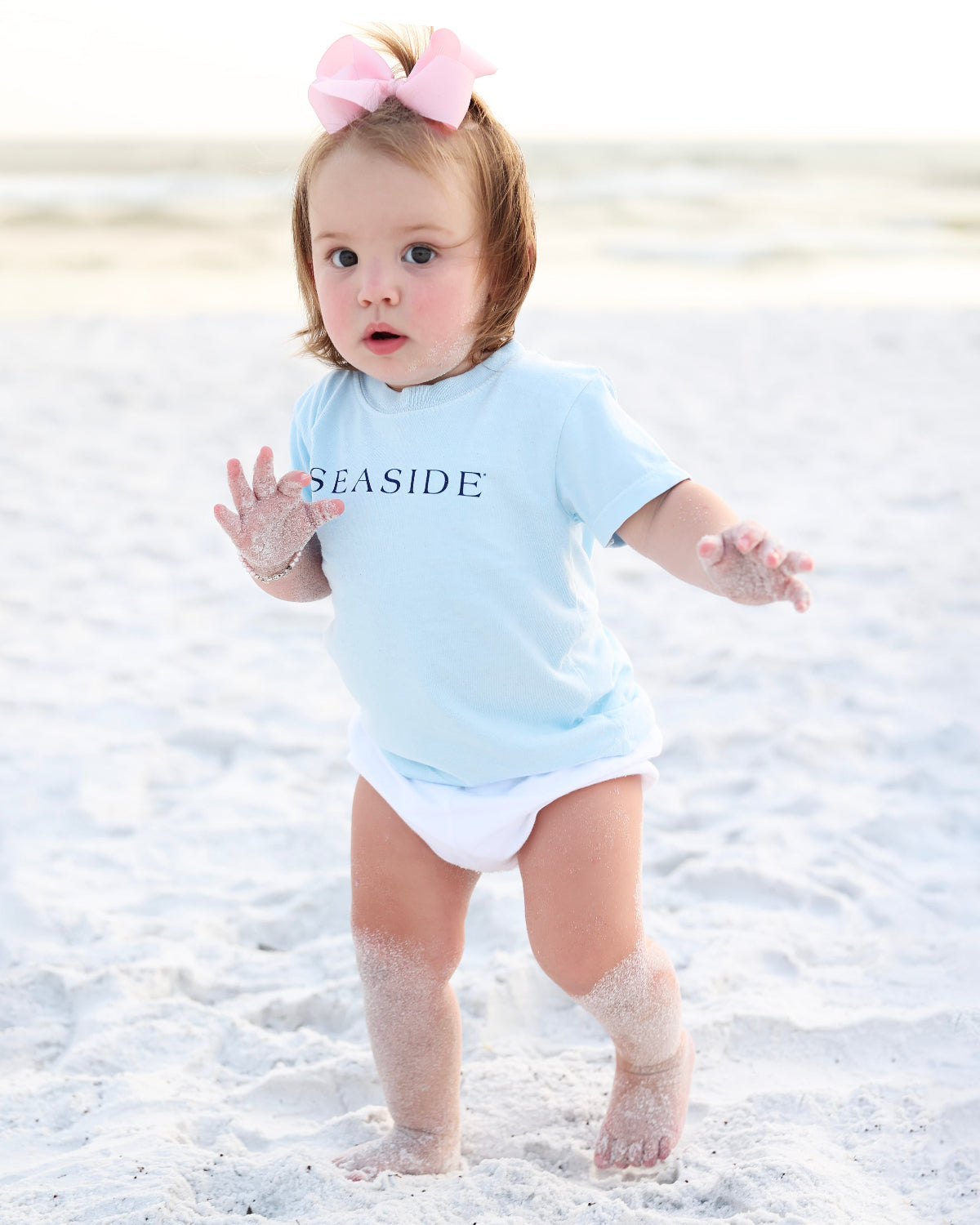 A toddler standing on a sandy beach wearing a light blue short-sleeve bodysuit with the word 'SEASIDE' printed in navy across the chest, along with a white diaper and a pink bow in her hair.