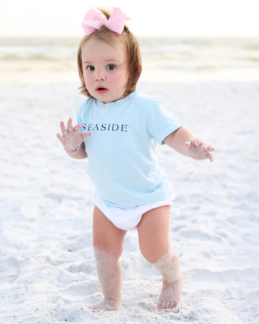 A toddler standing on a sandy beach wearing a light blue short-sleeve bodysuit with the word 'SEASIDE' printed in navy across the chest, along with a white diaper and a pink bow in her hair.