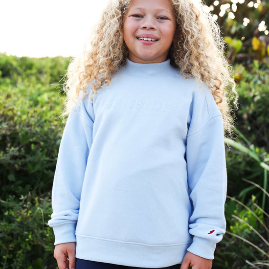 A smiling young girl with long curly blonde hair stands outdoors wearing a light blue mocknec sweatshirt with 'SEASIDE' printed across the chest and navy blue shorts, set against a background of green foliage.