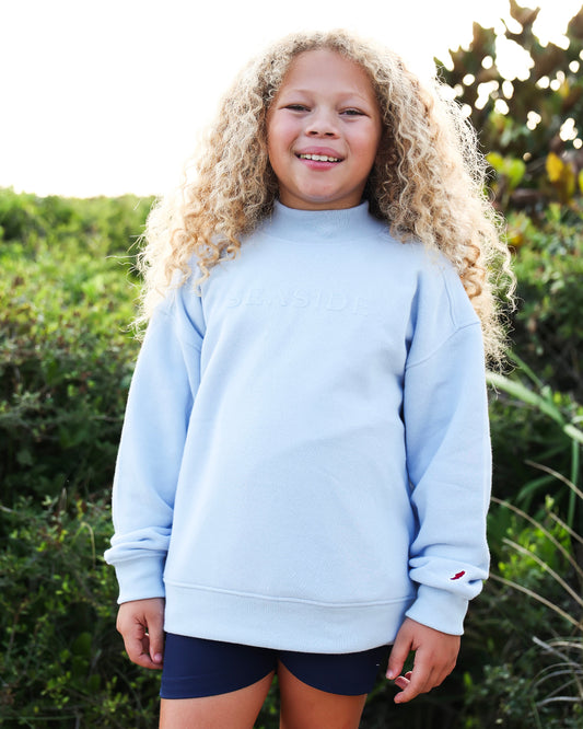 A smiling young girl with long curly blonde hair stands outdoors wearing a light blue mocknec sweatshirt with 'SEASIDE' printed across the chest and navy blue shorts, set against a background of green foliage.