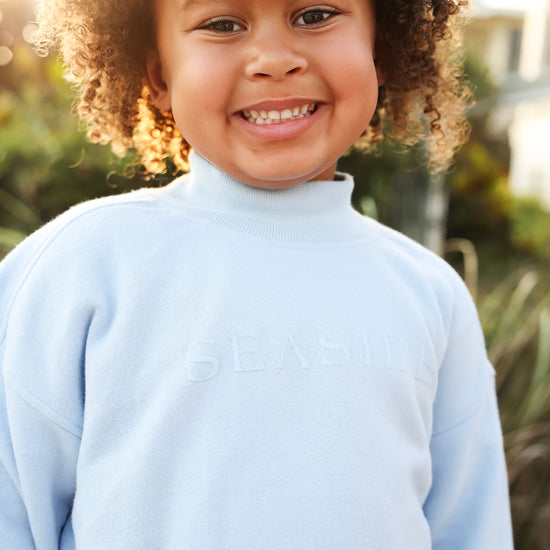 A smiling young child with curly hair wearing a light blue mock-neck sweatshirt with the 'SEASIDE' logo embossed on the chest, set against a warm, sunlit outdoor background.