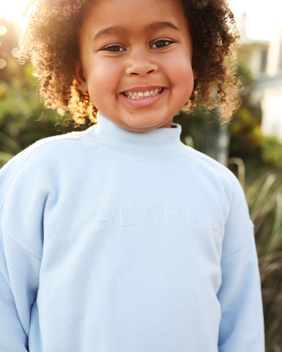 A smiling young child with curly hair wearing a light blue mock-neck sweatshirt with the 'SEASIDE' logo embossed on the chest, set against a warm, sunlit outdoor background.